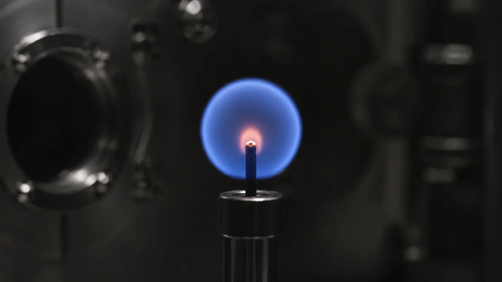 Chama azul redonda e pequena centralizada em um pavio, dentro de uma câmara escura de pesquisa.